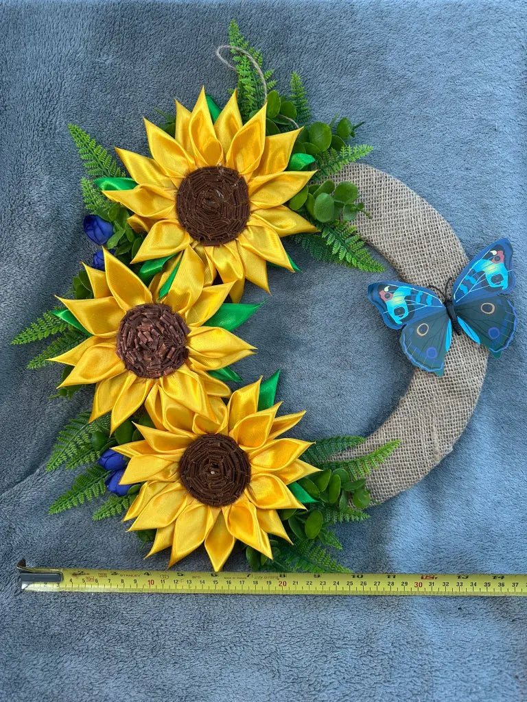 A decorative wreath with artificial sunflowers and a butterfly, placed against a blue background and measured with a tape measure for scale.