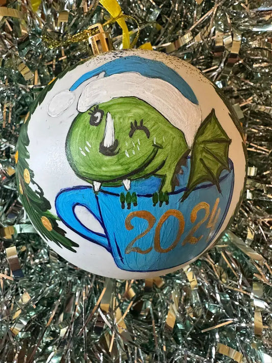 A hand painted Christmas bauble featuring a green figure in a blue cup with a white and gold '2024' patch on it, set against a tinsel background.
