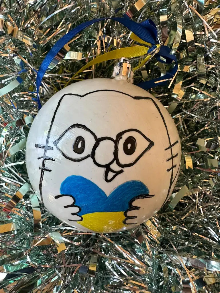 Hand painted Christmas bauble with a face and heart design, predominantly white with blue and yellow details, placed on a glittery surface.