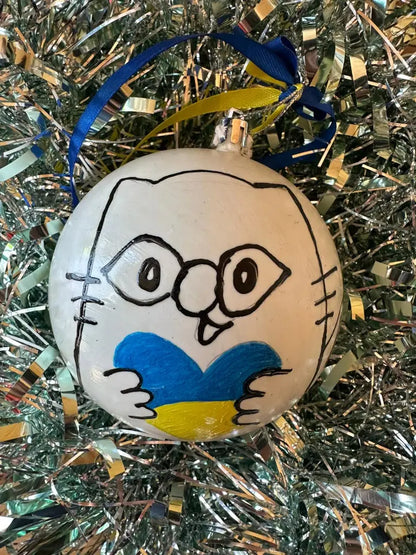 Hand painted Christmas bauble with a face and heart design, predominantly white with blue and yellow details, placed on a glittery surface.