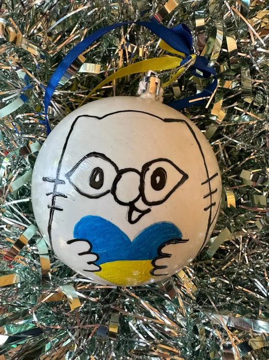 Hand painted Christmas bauble with a face and heart design, predominantly white with blue and yellow details, placed on a glittery surface.