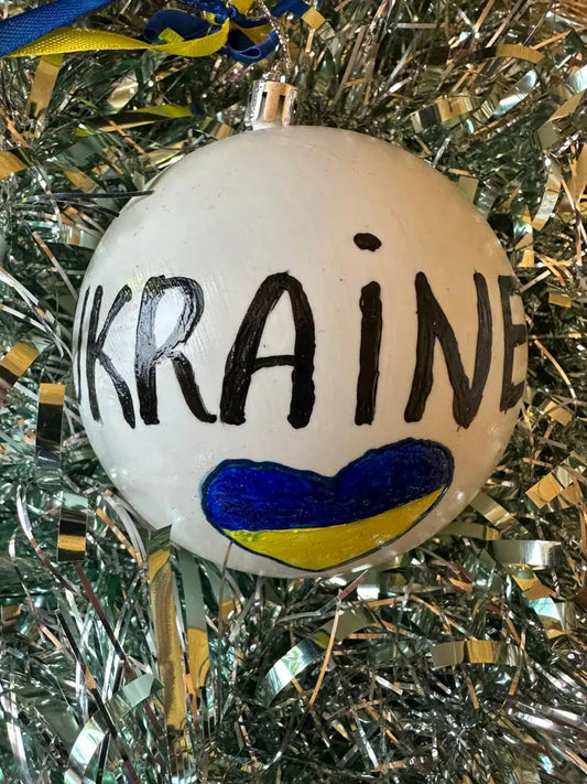 Hand painted white Christmas bauble with the word 'UKRAINE' in black text, decorated with a blue and yellow pattern at the top, set against a blue background with a yellow ribbon.