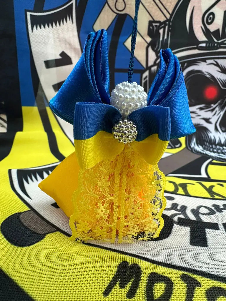 A handcrafted Christmas angel decoration featuring blue and yellow fabric with a white and yellow floral pattern, a lace crown, and a yellow sash, displayed against a backdrop with a yellow and black flag and pro-Ukrainian motifs.