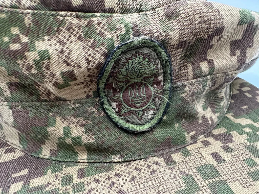 Camouflage military cap with a badge featuring a stylized emblem on a blue background