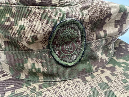 Camouflage military cap with a badge featuring a stylized emblem on a blue background